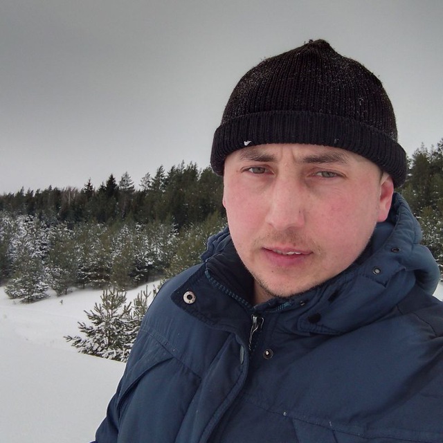 Фотография мужчины в черной шапке и синем пуховике. Он стоит на фоне зимнего леса и смотрит прямо.