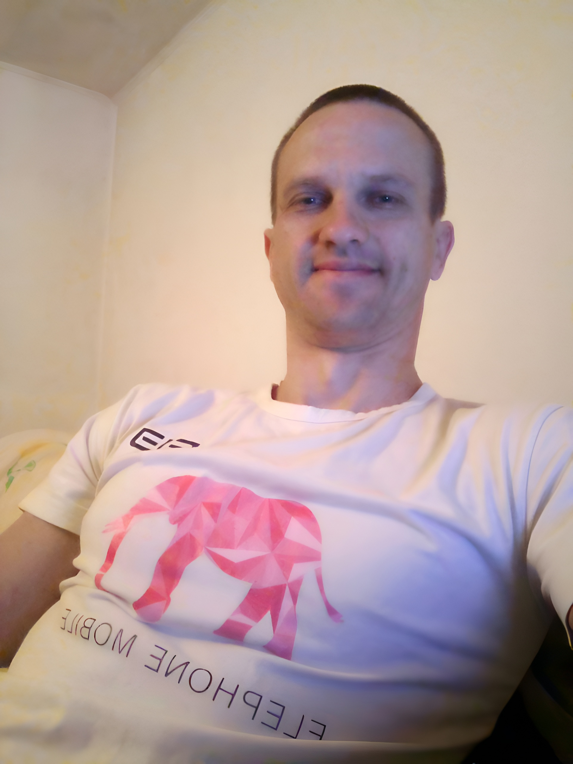 Молодой и красивый мужчина в белой футболке, на которой изображен розовый слон и надпись elephon mobile, сидит вполоборота на диване слегка улыбаясь и смотря прямо.